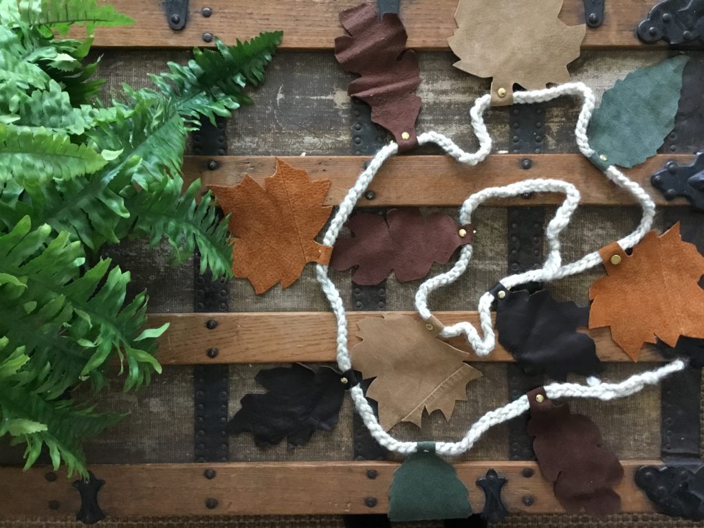 Fall leaves cut from scraps of leather and strung on a crocheted garland using brass fasteners, displayed on an old trunk with green fern