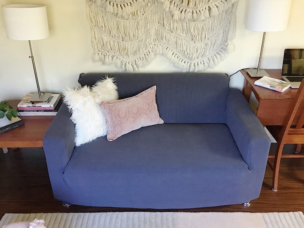 boho wall hanging above grey slipcovered sofa with fur pillow and rose denim pillow and ikea lamps and thrifted books on end tables