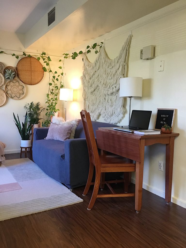 College campus housing with boho throw on wall, thrifted baskets on wall , slipcovered grey sofa ikea lamps and workstation in foreground