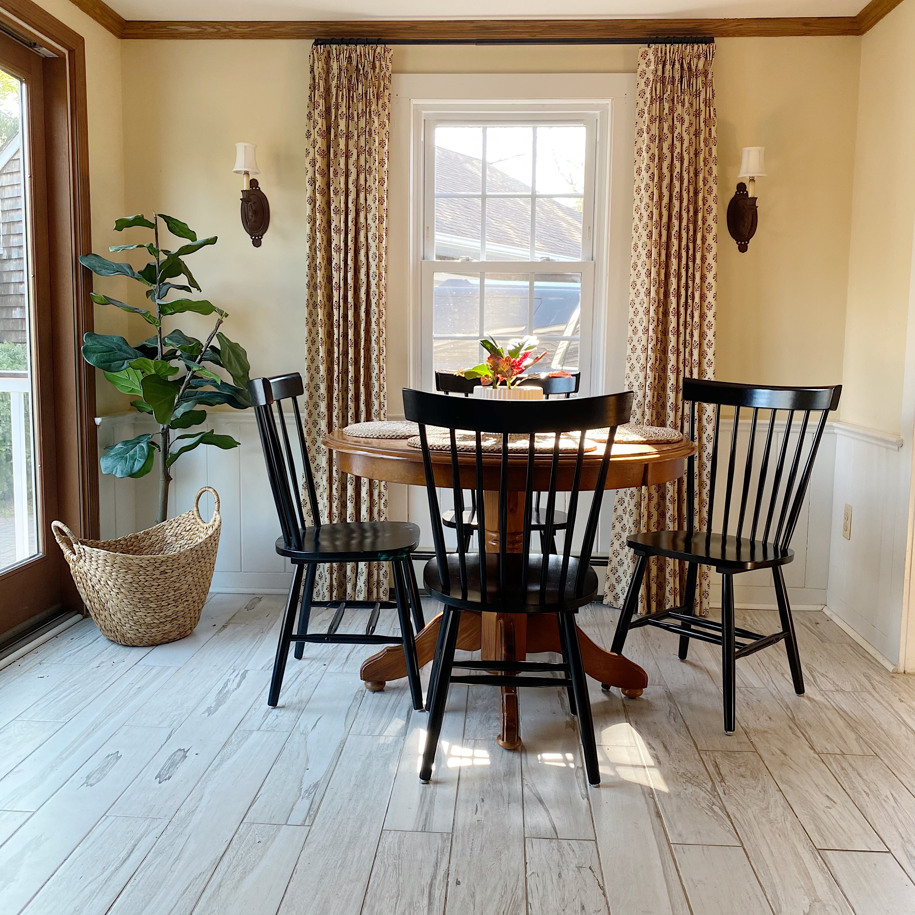 kitchen eating nook with vintage print drapes, pedestal table and black Windsor chairs with white wood tiles floor