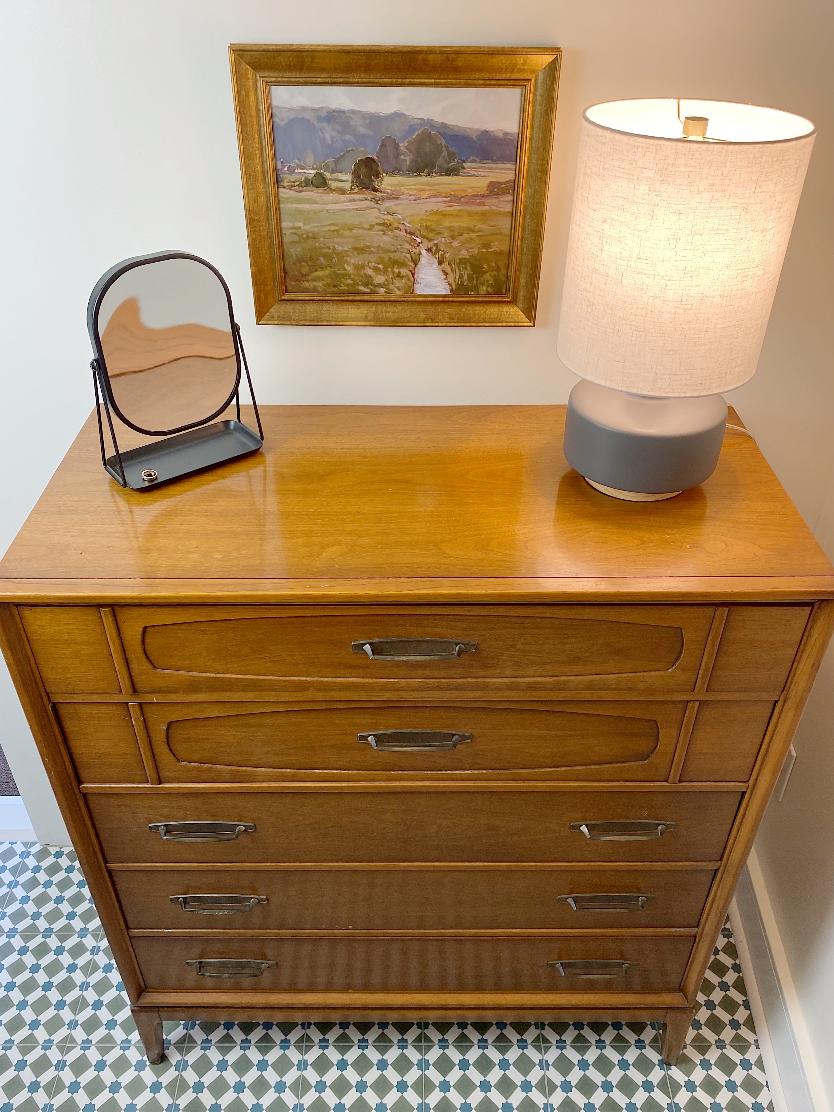 midcentury wood dresser in bathroom with small lamp and wall print and patterned floor tile