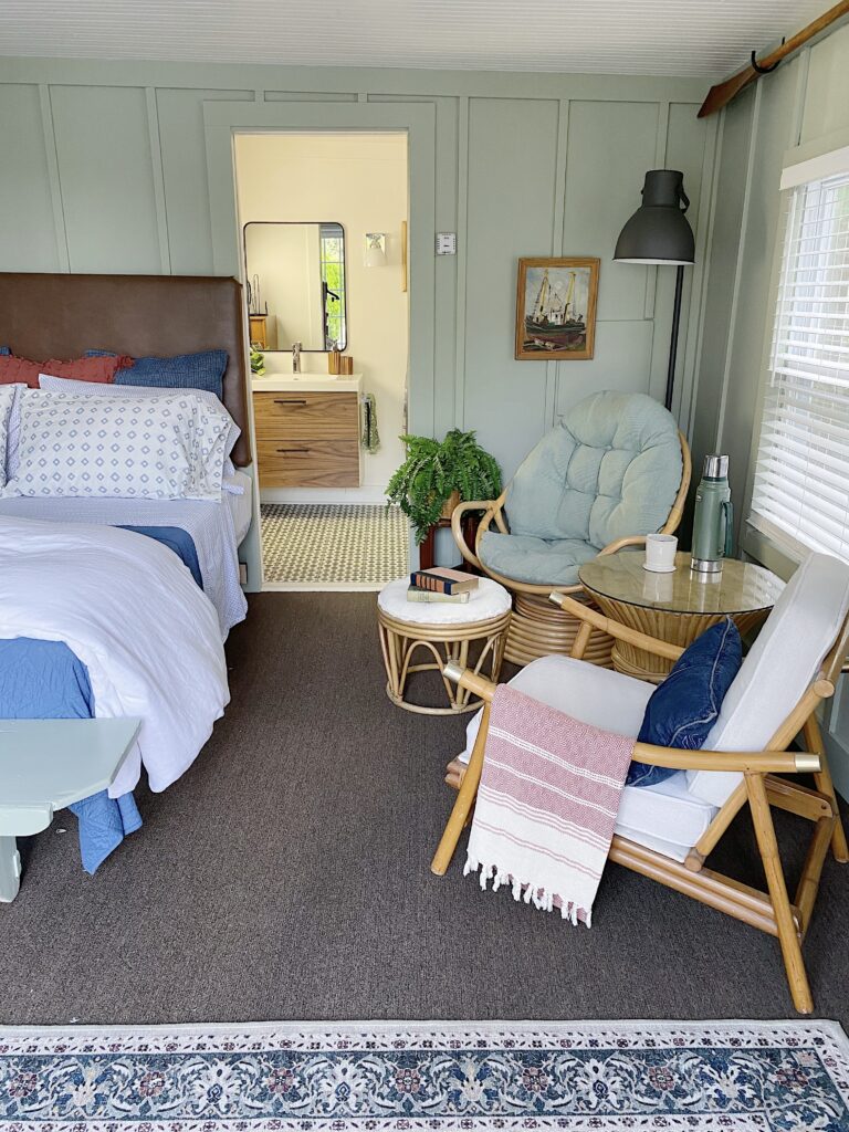 small guest room with leather bed, linen duvet and small print sheets and sitting area with vintage rattan chairs, green board and batten walls